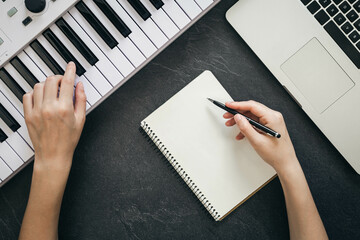 Female hands, blank notepad, piano and laptop on a black background, top view.