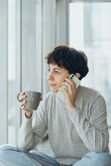 A woman with a short haircut talks on the phone and drinks tea in the morning.