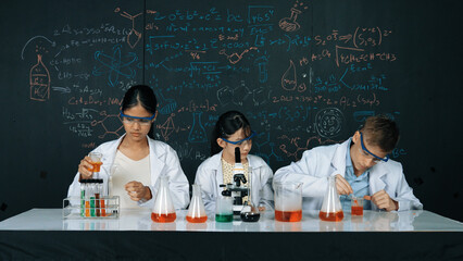 Cute girl looking under microscope while student doing experiment at blackboard with theory written. Young scientist inspect colored solution at table with experimental equipment placed. Edification.