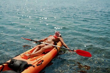 A woman is laying on a kayak in the water. The kayak is orange and has a paddle on it. The woman is...
