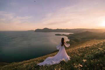A woman in a white dress stands on a hill overlooking the ocean