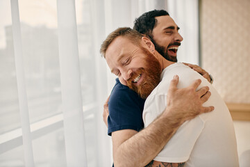 Two men in casual clothes embrace each other with love in a modern living room, captured in a tender moment by the window.
