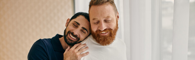 Two men in casual attire hug affectionately in front of a large window, embodying love and...