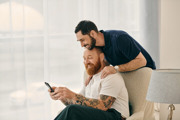 Casual gay couple lounging in a chair, engrossed in cell phone, sharing a moment of modern...