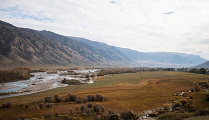 Landscape during autumn. In the foreground, grassland and meandering river bordered by trees with autumnal foliage and field with sparse vegetation. Mountains obscured by haze on background