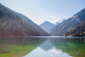 a serene alpine lake with crystal-clear waters reflecting the sky, bordered by dense coniferous...