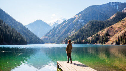 a adult woman standing on a dock overlooking a tranquil alpine lake, gazing at the snow-covered mountain peaks and surrounding dense pine forests under a clear blue sky - Powered by Adobe
