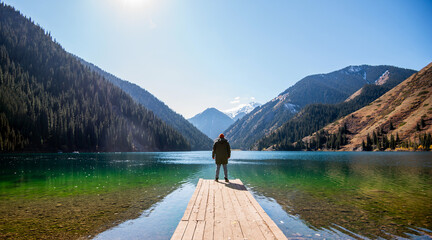 a man standing at the end of a wooden pier, looking out over a clear alpine lake towards towering,...