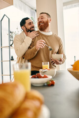 Two men hug in the kitchen while enjoying breakfast together in a modern apartment, expressing...