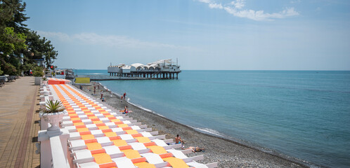 A sunny pebble beach with colorful sun loungers and a pier hosting a white pavilion, set against a...