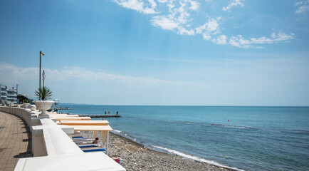 A serene pebble beach with sunbeds lined along a promenade, a clear blue sky above, and a calm sea...