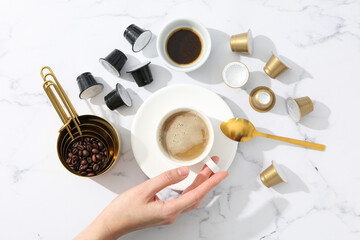 Coffee capsules and beans, cups with coffee drink and hand on marble background, top view