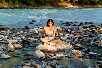 Young fit beautiful woman sitting on stone in mountain river playing with water on nature in...