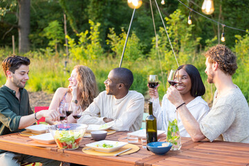 A group of friends is having a delightful dinner party outdoors, sharing wine, food, and engaging...