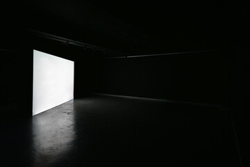 Big blank white illuminated screen backdrop in large empty dark room concrete floor. Nobody in room.