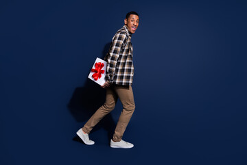 Full length photo of good mood guy wear checkered shirt walking hiding gift box emtpy space isolated blue color background