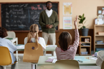 Selective focus shot of unrecognizable boy sitting at desk in classroom raising hand in English...