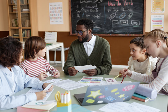 Young African American male teacher of English sitting at table in classroom working with multi-ethnic group of kids