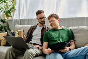 Two men in love sit on a couch, focused on a laptop, designing trendy attire in their workshop.