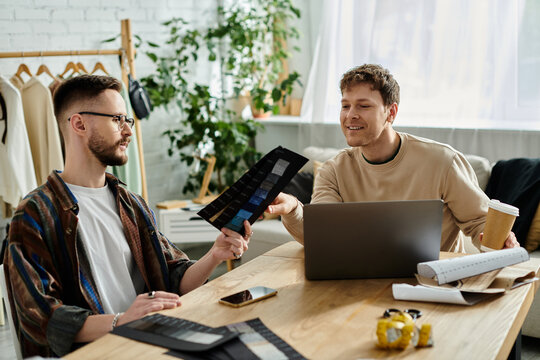 Two men, a gay couple, sit at a table with a laptop, collaborating on trendy attire they design together in their workshop. - Powered by Adobe
