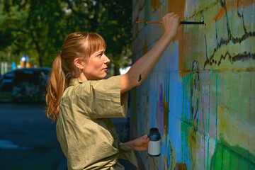 Pretty graffiti artist painter with a brush on a stepladder paints a wall