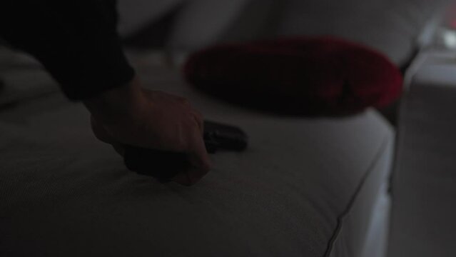 An obscured man's hand reaches for a handgun on a couch in a dimly lit, tense indoor setting.