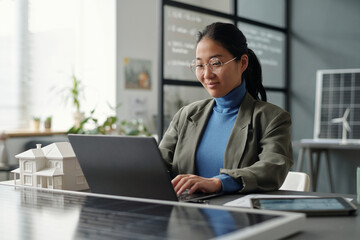 Smiling Asian female employee looking at laptop screen and typing while sitting by workplace with house model and solar panel