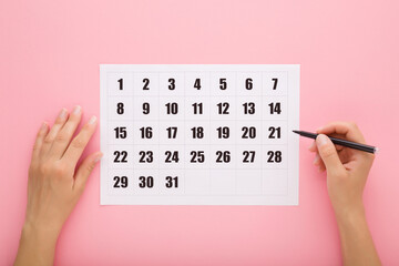Young adult woman hand holding dark black color pen and marking day in white calendar on light pink...