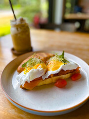 Close-up of gourmet poached eggs topped with hollandaise sauce on smoked salmon and a toasted baguette, served with cherry tomatoes and iced coffee in a cozy cafe. Ideal for food blogs, culinary