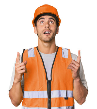 Young Hispanic worker in safety gear pointing upside with opened mouth.