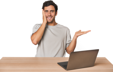Hispanic young man working on laptop holds copy space on a palm, keep hand over cheek. Amazed and delighted.
