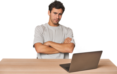 Hispanic young man working on laptop suspicious, uncertain, examining you.