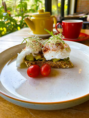 Close-up of a healthy avocado toast topped with poached eggs and sprouts, served with cherry tomatoes and a tea set in a cozy cafe. Perfect for health-conscious food blogs, breakfast menus, and