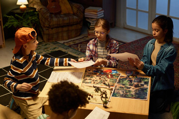 Four cute intercultural children sitting on the floor by table with boards, cards, dice and other...