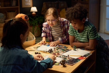 Group of intercultural tense children bending over table with leisure game board with characters...