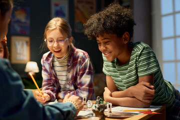 Cheerful kids standing by table with new board game, looking at dice thrown by one of boys and laughing while enjoying play