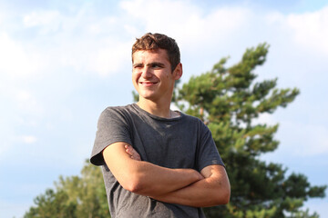 Young smiling man on the summer day. Happy and confident man with crossed arms. Candid young man outside. Nature blurred background