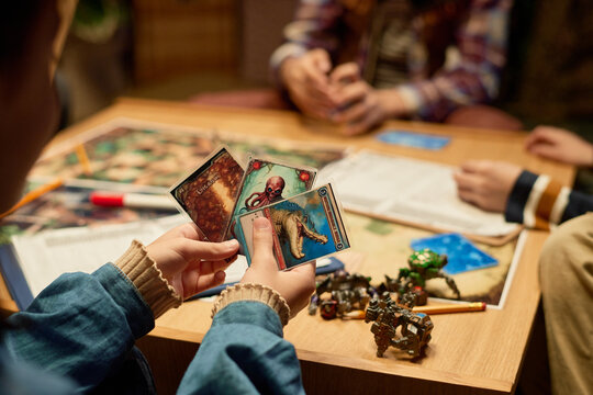 Hands of unrecognizable girl in denim jacket holding three board game cards and choosing one of them while playing with her friends