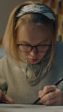 Focused illustrator draws sketches for her project. Young female artist with glasses dressed in a grey hoodie and sits on her work desk. Makes pencil sketches. Story telling concept. Vertical shot.