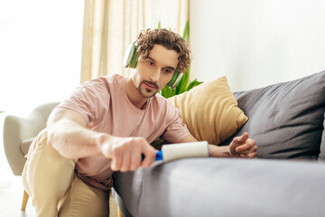 Handsome man in cozy homewear sitting on couch with sticky roller in hand.