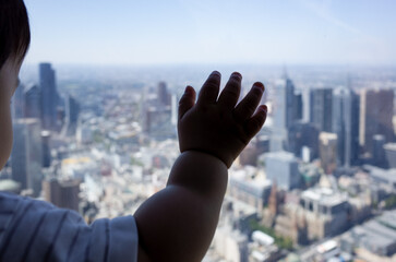 Close-up of a 9-month-old Asian baby's hand touching a glass window with a blurry elevated complex...