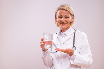 Portrait of mature female doctor showing glass of water  on gray background.