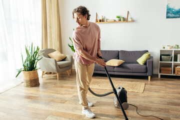 A handsome man in cozy homewear uses a vacuum to clean his living room.