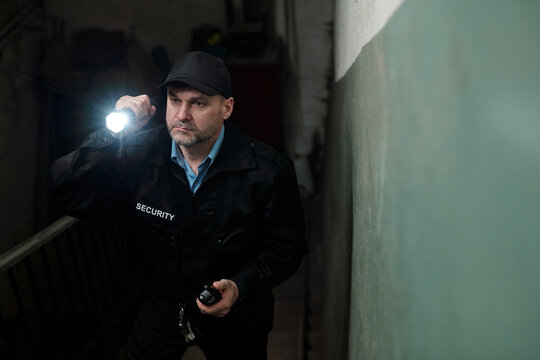 Serious mature guard in uniform moving upstairs along narrow corridor in darkness and lighting his way with flashlight during night shift - Powered by Adobe