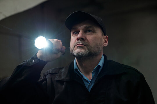 Mature security guard with flashlight observing dark corridor or office while directing light forwards during work at night