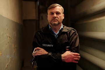Serious mature male security guard with bunch of keys in hand crossing arms by chest and looking at...