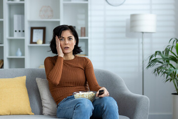 Scared and worried young woman sitting on sofa at home, holding bowl of popcorn on lap and looking ahead with fear, watching scary movie and news, holding hand to head in shock