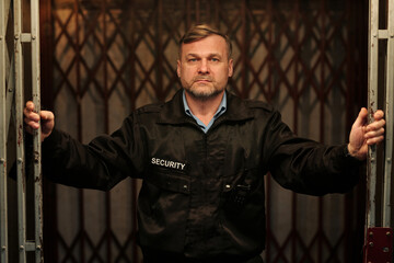 Serious security guard in black uniform looking at camera while opening or holding mesh gate in the...