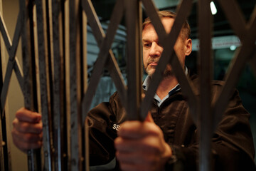 Serious mature male guard standing behind mesh and opening gate before entering one of rooms of...