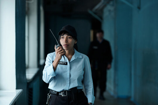 Young female security guard in uniform and cap speaking on walkie-talkie to her colleague while moving along corridor of building